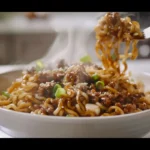 A bowl of Mongolian ground beef noodles with green onions and sesame seeds on a white marble background.